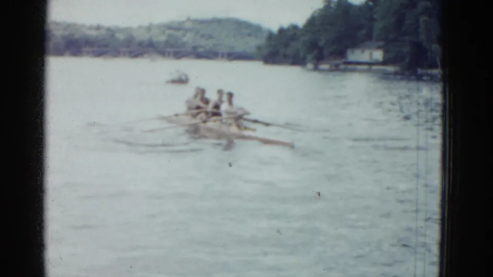 Soft-focus 1975 home-movie frame of a four-man rowing shell moving away on a calm lake near a wooded shoreline.