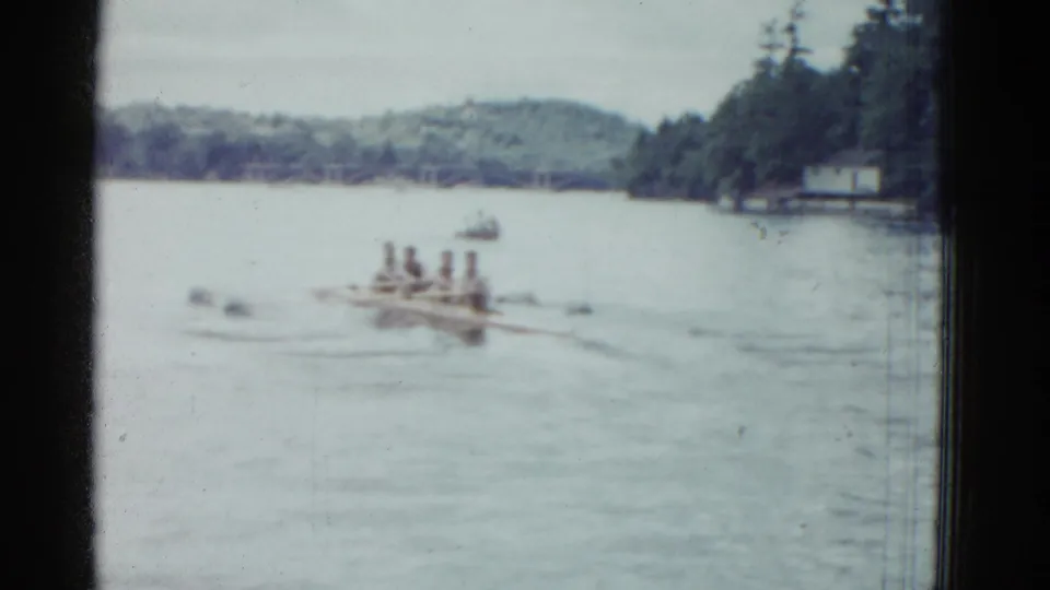 A 1975 Boston-area 8mm frame shows the rowers near mid-lake, with a distant small boat, pine-lined shore, and overcast sky.