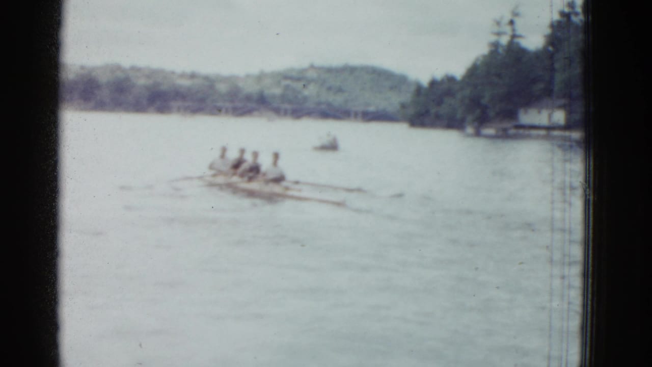 Four rowers in a shell move across a lake with oars extended and wooded shoreline behind them.