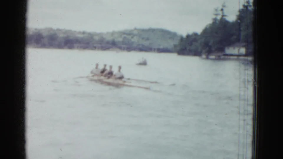 1975 Boston-area 8mm frame showing four rowers in a narrow shell crossing a calm lake near a wooded shore.