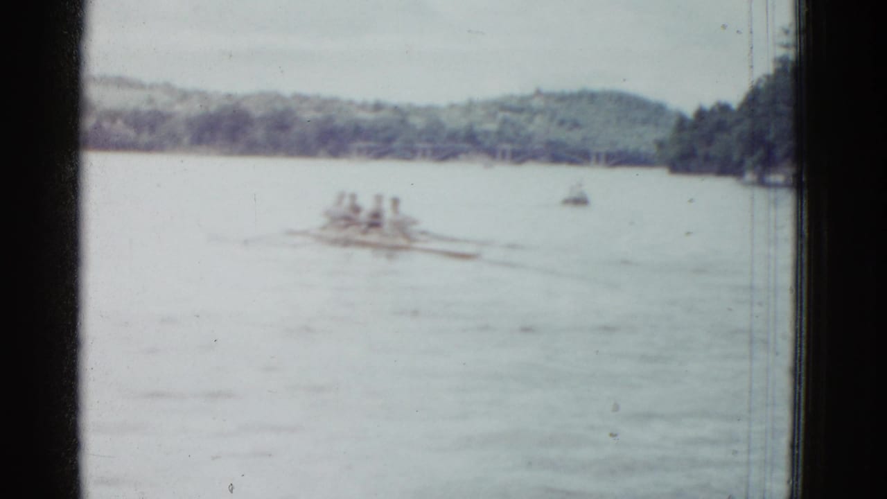 1975 8mm home-movie frame of rowers moving across a lake, with soft focus, a small distant boat, and trees along the shore.