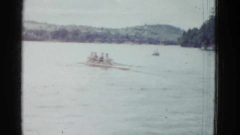 1975 8mm home-movie frame of rowers moving across a lake, with soft focus, a small distant boat, and trees along the shore.