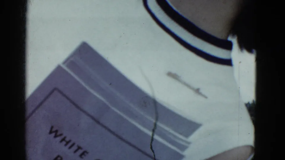 Blurry 1975 home-movie transition showing part of a person in a white shirt near a vehicle or equipment, with film scratches visible.