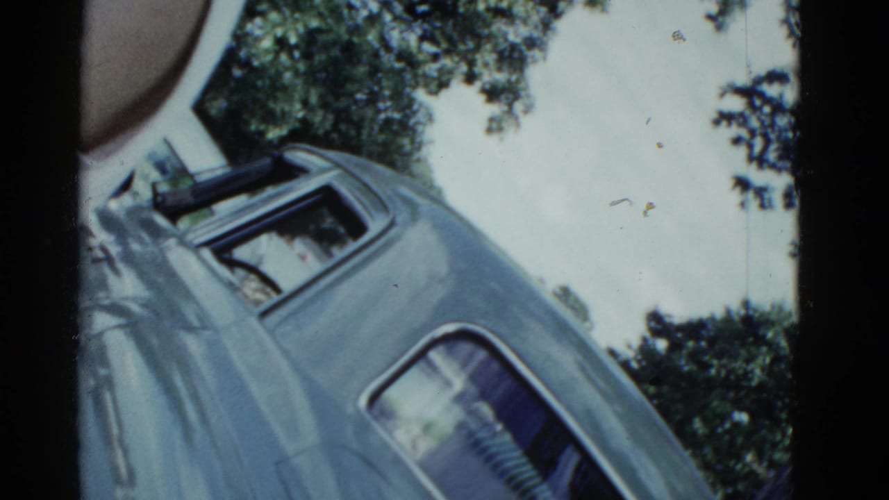 1975 Boston 8mm frame showing a tilted view of a blue car roof and windows against bright sky and leafy trees, with a partial person at left.