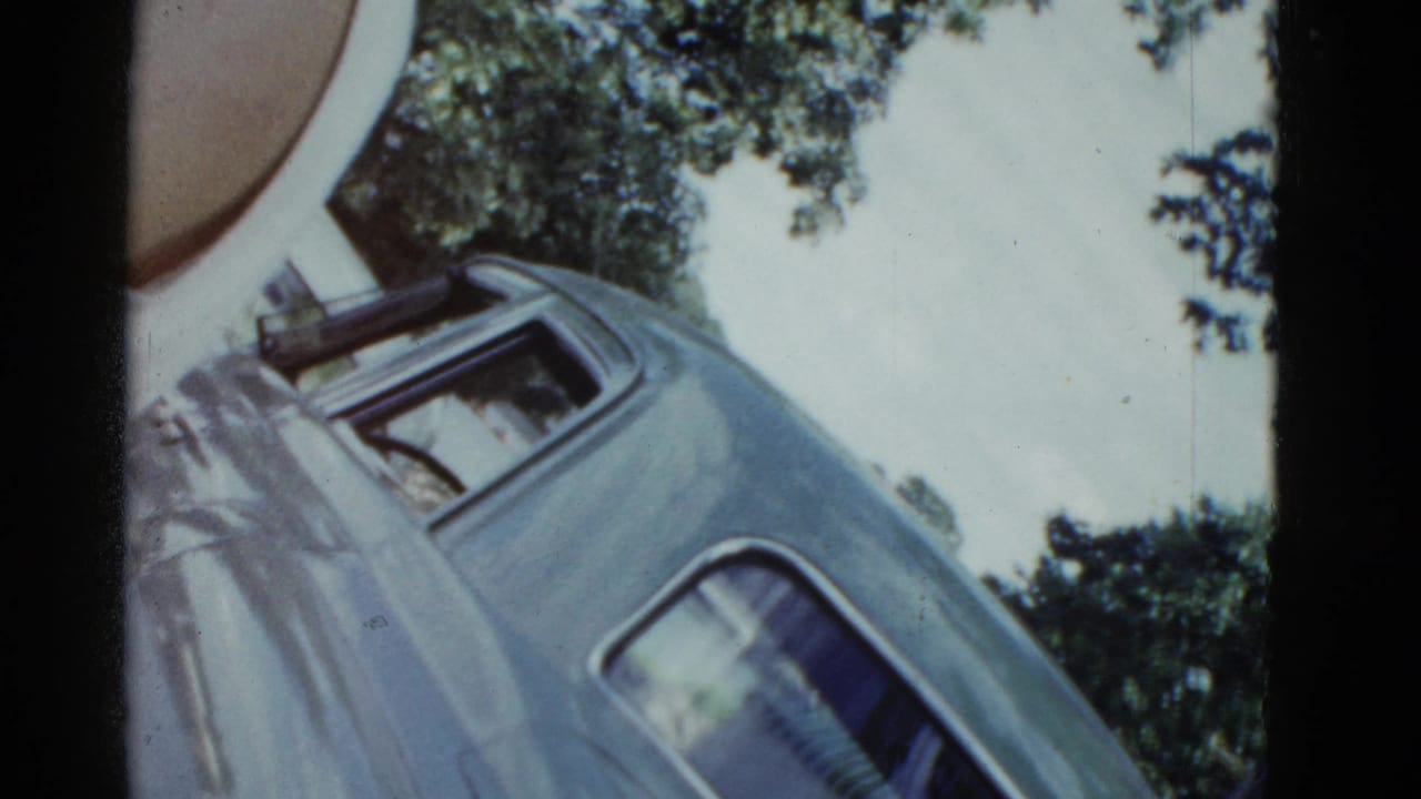 1975 Boston home-movie frame with a blue car angled across the view, tree canopy above, and a partial person near the left edge.