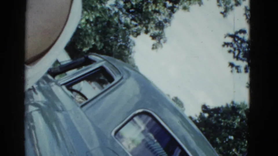 1975 Boston 8mm still showing the blue car from a low tilted angle, with leafy trees, pale sky, and a partial foreground person.