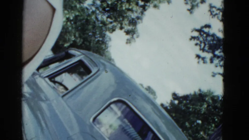 1975 Boston frame of a blue car shot from below at a diagonal, with sky, tree leaves, film edges, and a partial person at left.