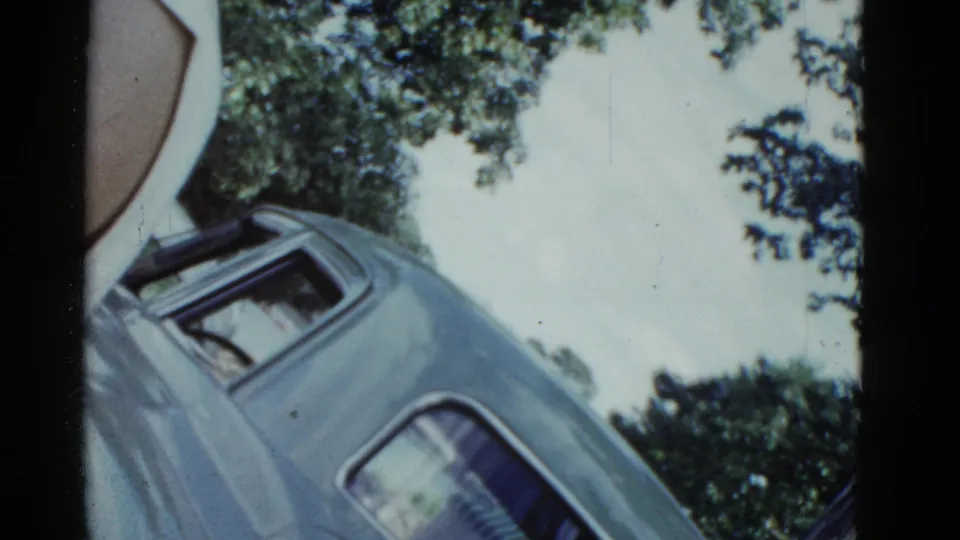 1975 8mm home-movie frame from Boston area showing a tilted parked car near leafy trees and waterfront light.