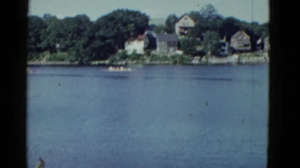 1975 Boston lakeside view with blue water, distant houses, trees, and a small boat crossing the frame.