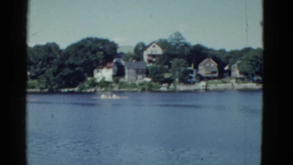 Handheld 8mm frame of a Boston lakeside scene with a rowboat on open water and residential buildings beyond the shore.