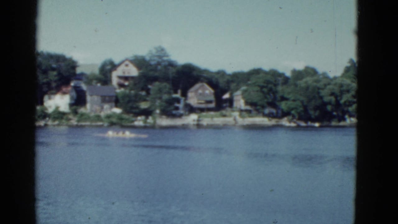 Two rowing shells cross blue water with houses and trees on the far shore.