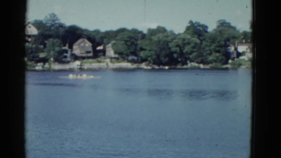 Soft 8mm frame from the 1975 Boston clip showing rowers on calm water near a leafy residential shore.