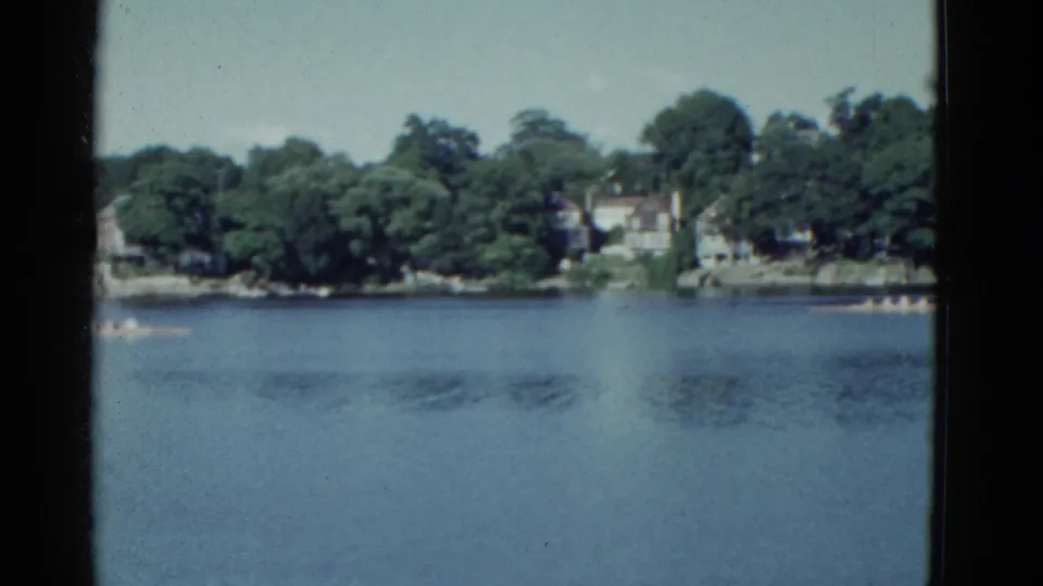 1975 Boston-area 8mm frame showing racing shells near the edges of calm water, with dense trees and houses beyond.