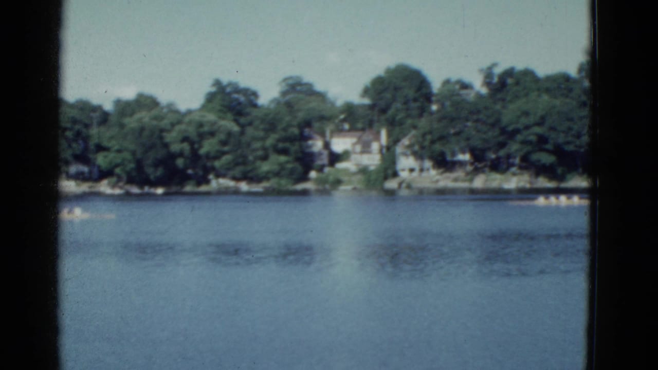 1975 Boston-area 8mm frame showing distant rowers on a calm river below a leafy residential shoreline.