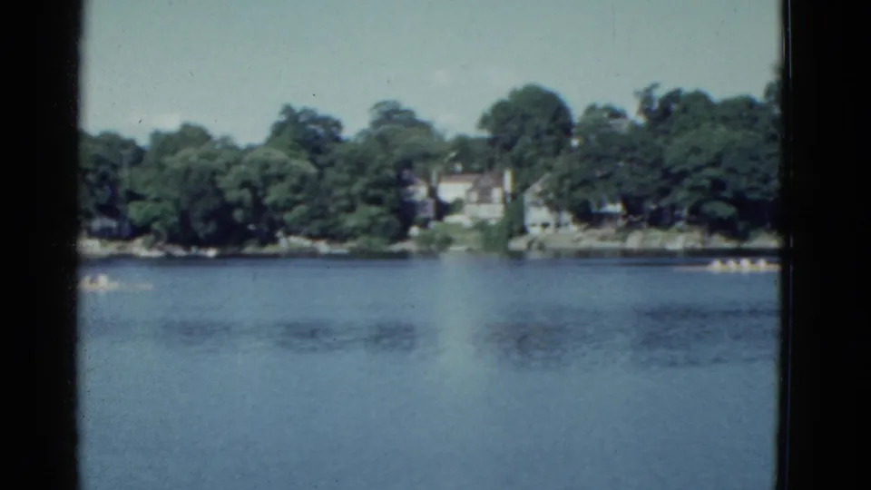 1975 Boston-area 8mm frame showing distant rowers on a calm river below a leafy residential shoreline.