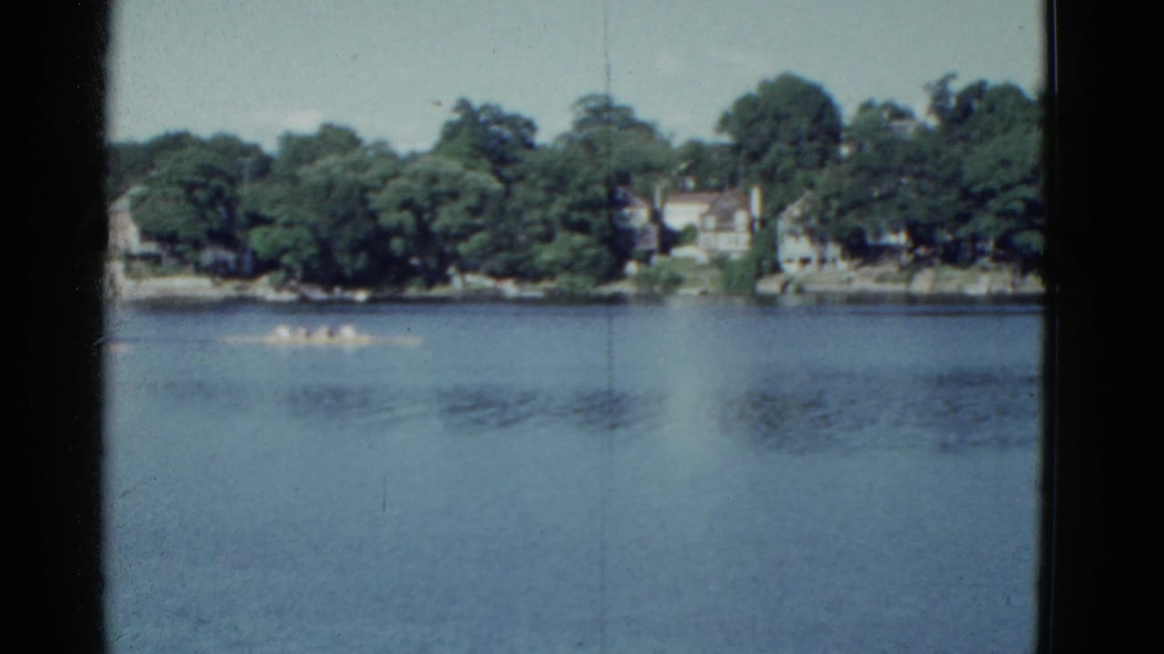 Wide 8mm view of a rowing shell on the water, with Boston-area trees and distant homes beyond the river.