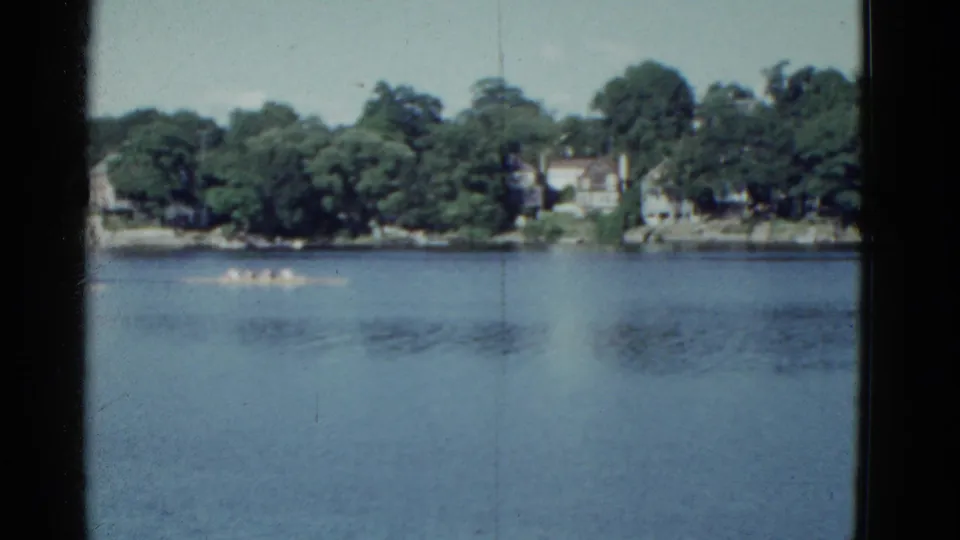Wide 8mm view of a rowing shell on the water, with Boston-area trees and distant homes beyond the river.