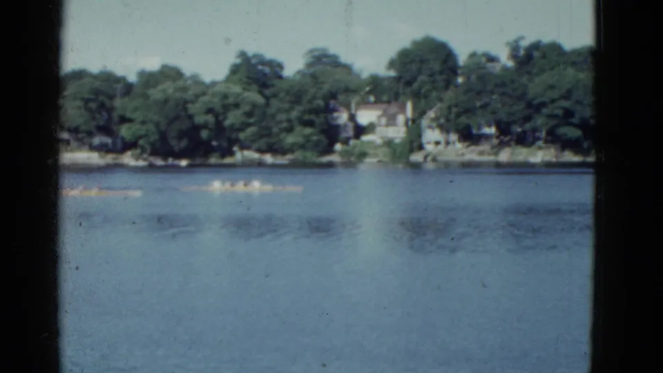1975 home-movie frame showing multiple distant rowing shells on a calm Boston-area river.