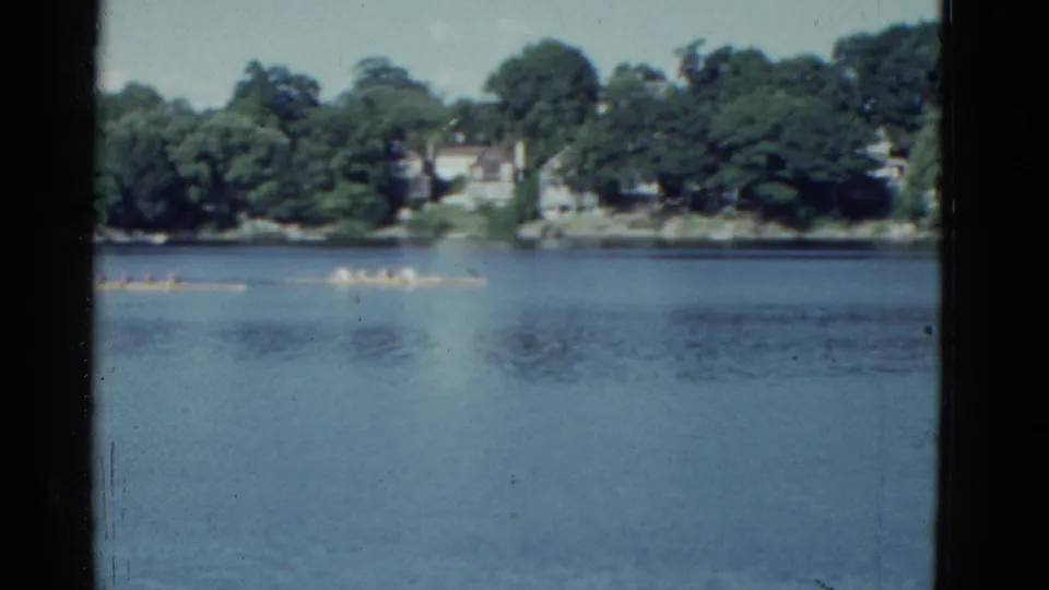 Soft 8mm frame of distant rowers moving across a calm river near Boston in 1975.