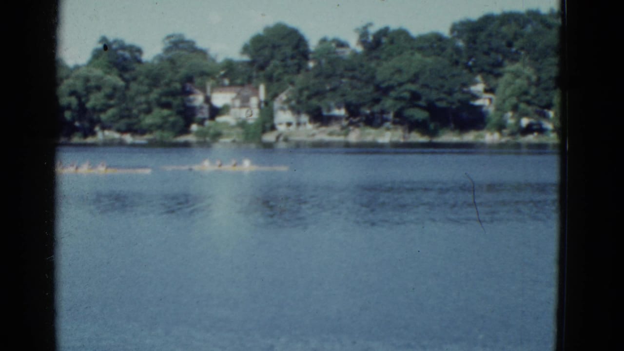 1975 Boston-area 8mm frame showing distant rowing crews crossing blue water below a tree-lined residential shoreline.