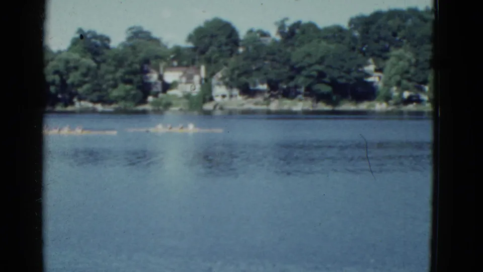 1975 Boston-area 8mm frame showing distant rowing crews crossing blue water below a tree-lined residential shoreline.