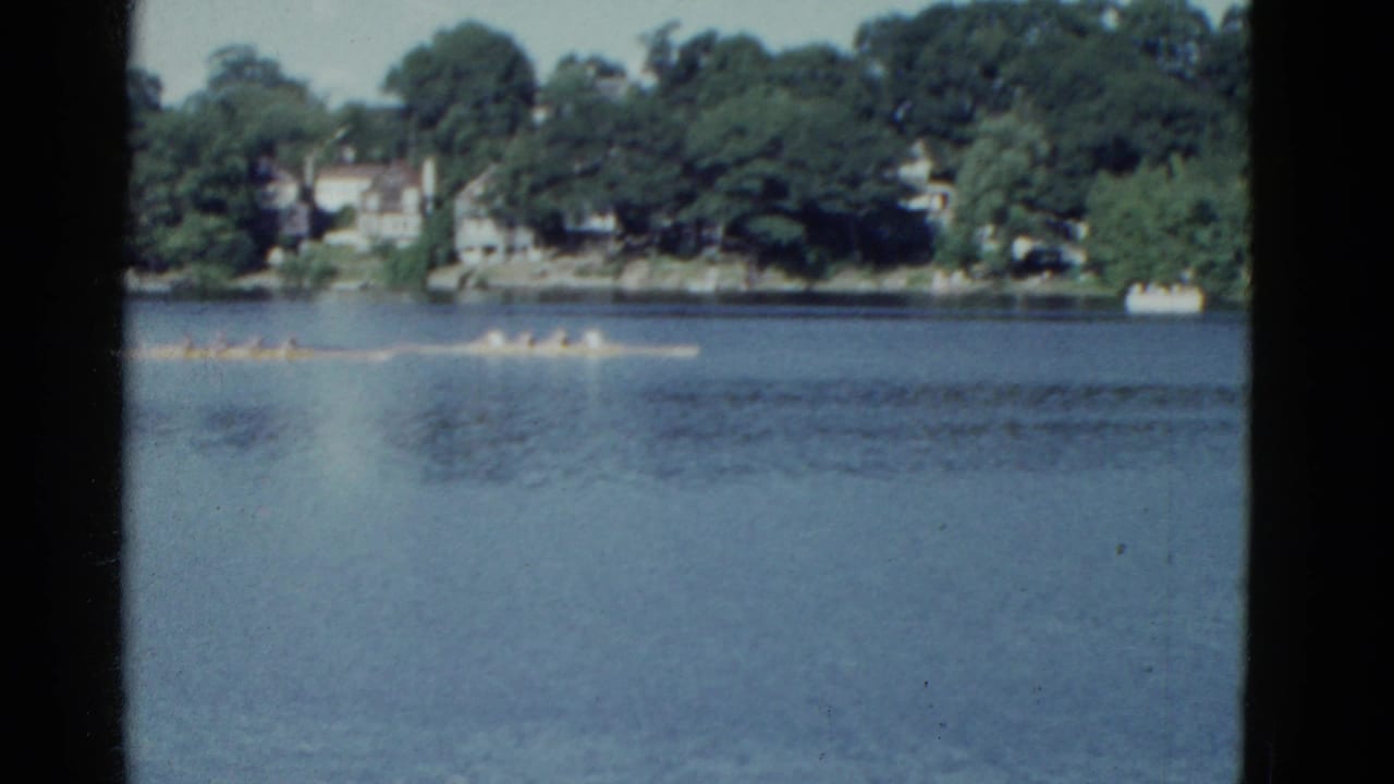 Distant 1975 rowing action on a calm Boston-area lake, with houses and dense trees visible beyond the water.