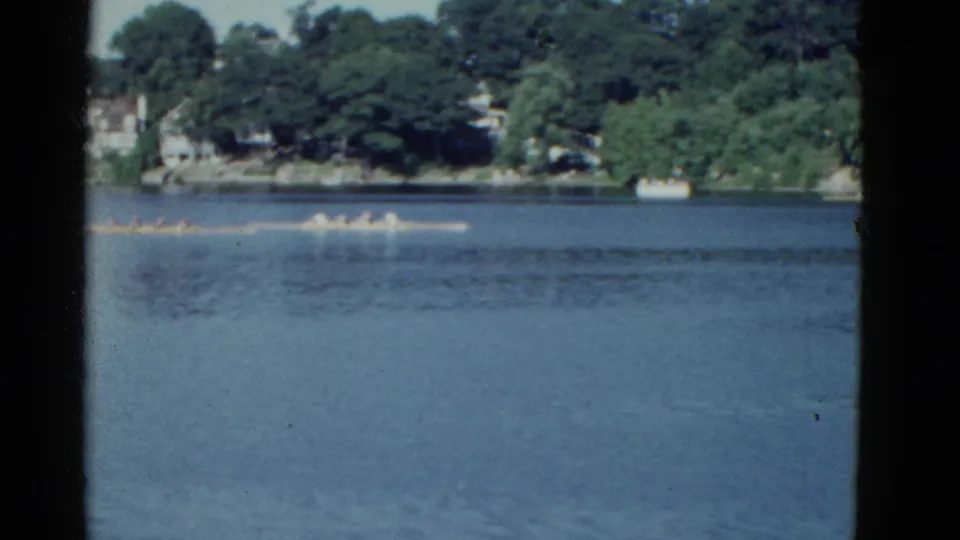 Soft 8mm frame of rowers moving across a Boston-area lake in 1975, with trees, homes, and a small shore boat in the background.