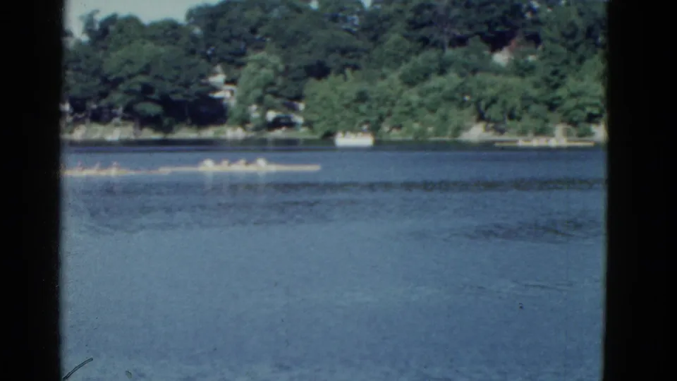 1975 8mm view of rowing crews on calm water near Boston, framed by dark film edges and a residential tree-lined shore.