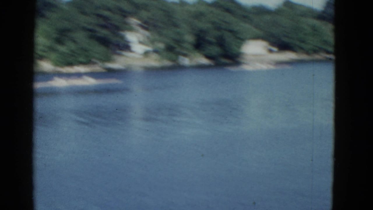 1975 Boston-area 8mm frame showing calm water, wooded shore, and a distant rowing shell near the left side of the view.