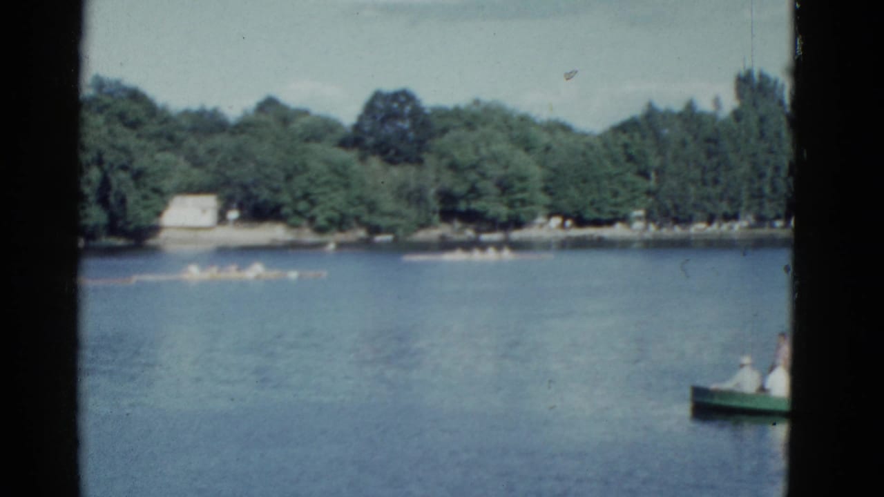 Green rowboat on a lake with a distant scull crew and tree-lined shore.