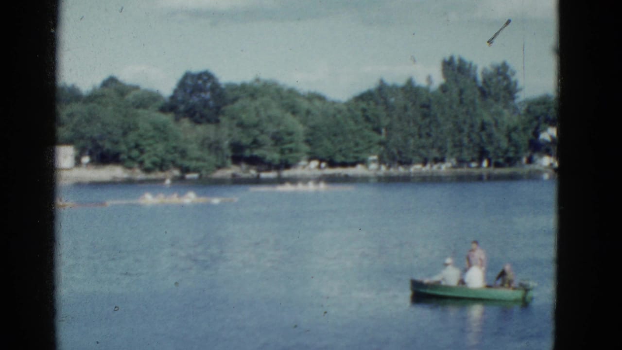 1975 Boston home-movie frame with people in a green rowboat at right and a long scull crew crossing the lake in the background.