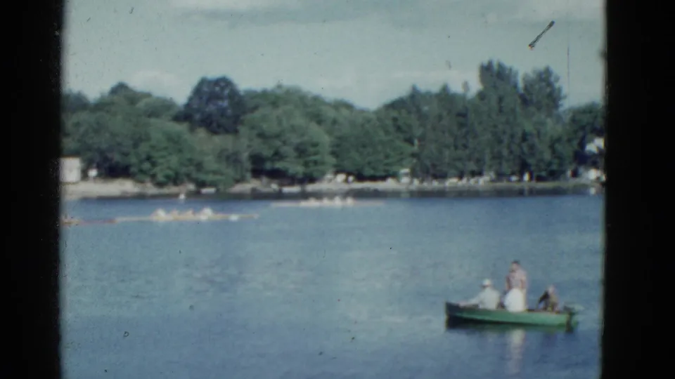 1975 Boston home-movie frame with people in a green rowboat at right and a long scull crew crossing the lake in the background.