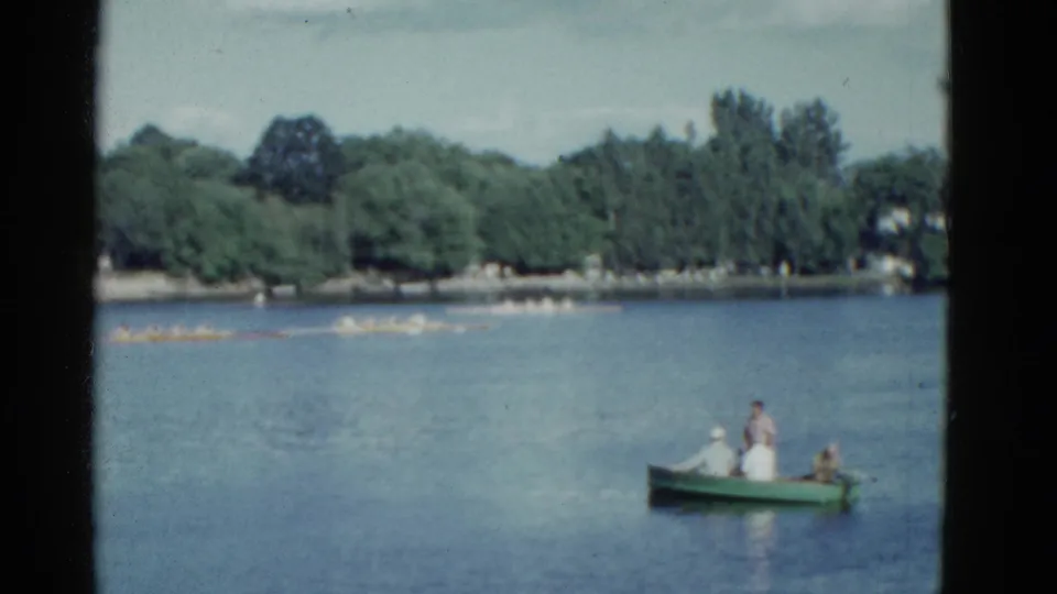 1975 Boston 8mm frame showing a small green rowboat in the lower right and a scull crew moving along the tree-lined far shore.