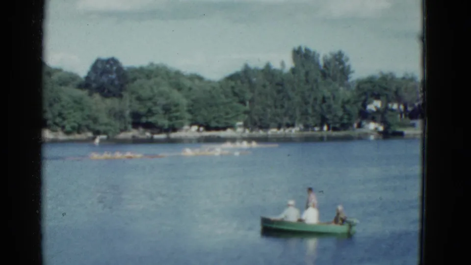 1975 Boston home-movie frame with a green spectator rowboat in the foreground and a rowing shell stretched across the distant lake.