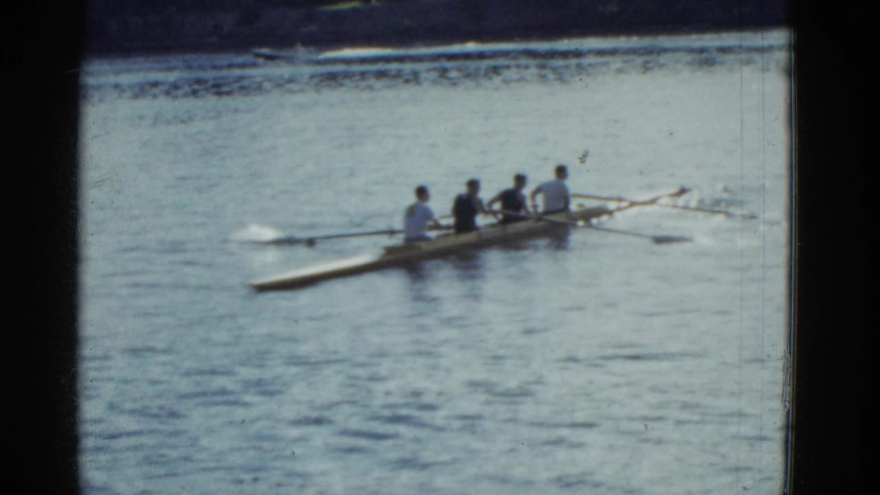 Four rowers in a long shell move across calm lake water.