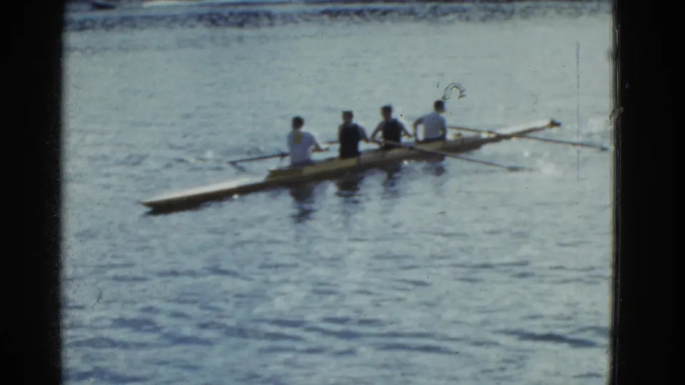 A four-person rowing shell moves across blue water in 1975 Boston-area home-movie footage, with the rowers seen from behind.