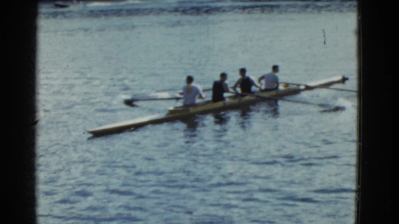 The rowing shell continues across the lake, with four visible rowers and wide oars in 1975 8mm footage.