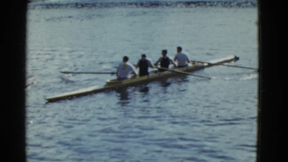From a slightly rearward angle, four young men row a narrow shell on a lake in 1975 Boston-area footage.