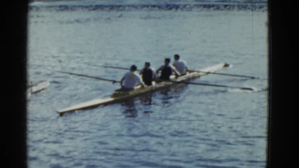 The four-person shell angles across the lake, showing synchronized oars and soft 8mm home-movie texture.