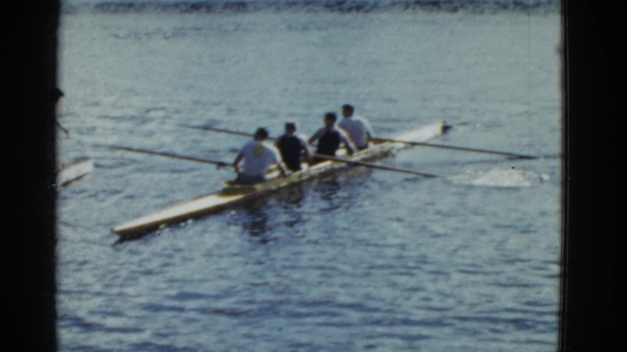 Four rowers in a narrow shell move across calm water near the edge of a dock.