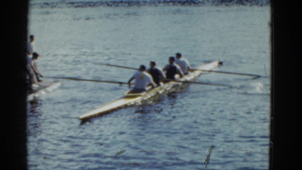 1975 Boston home-movie frame of a four-person rowing shell near dockside onlookers, filmed with soft grain and visible gate edges.