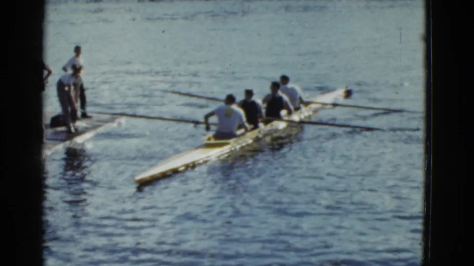 1975 Boston 8mm frame showing a four-person crew rowing beside a dock, with onlookers at left and oars spread over the water.