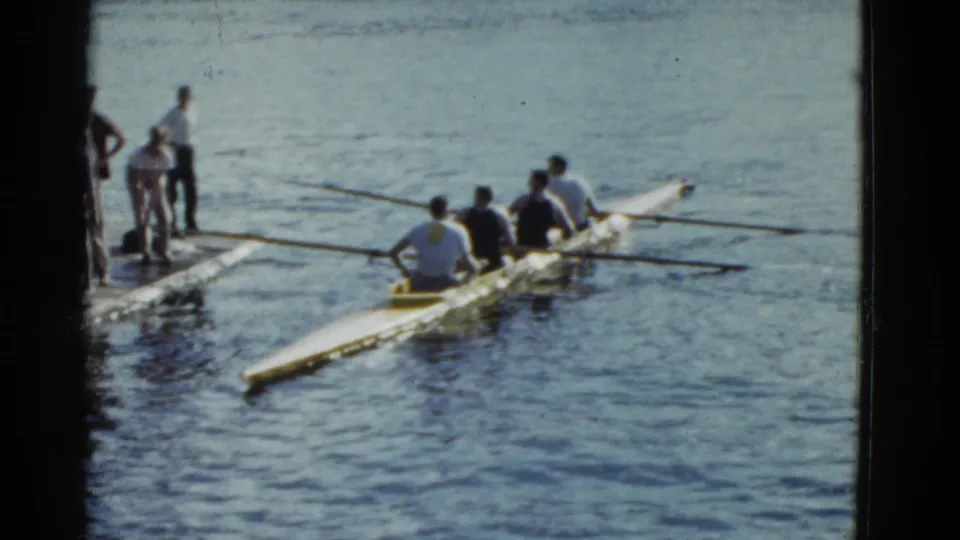1975 Boston home-movie frame of men rowing in a shell near a dock, with casual observers and visible 8mm film wear.