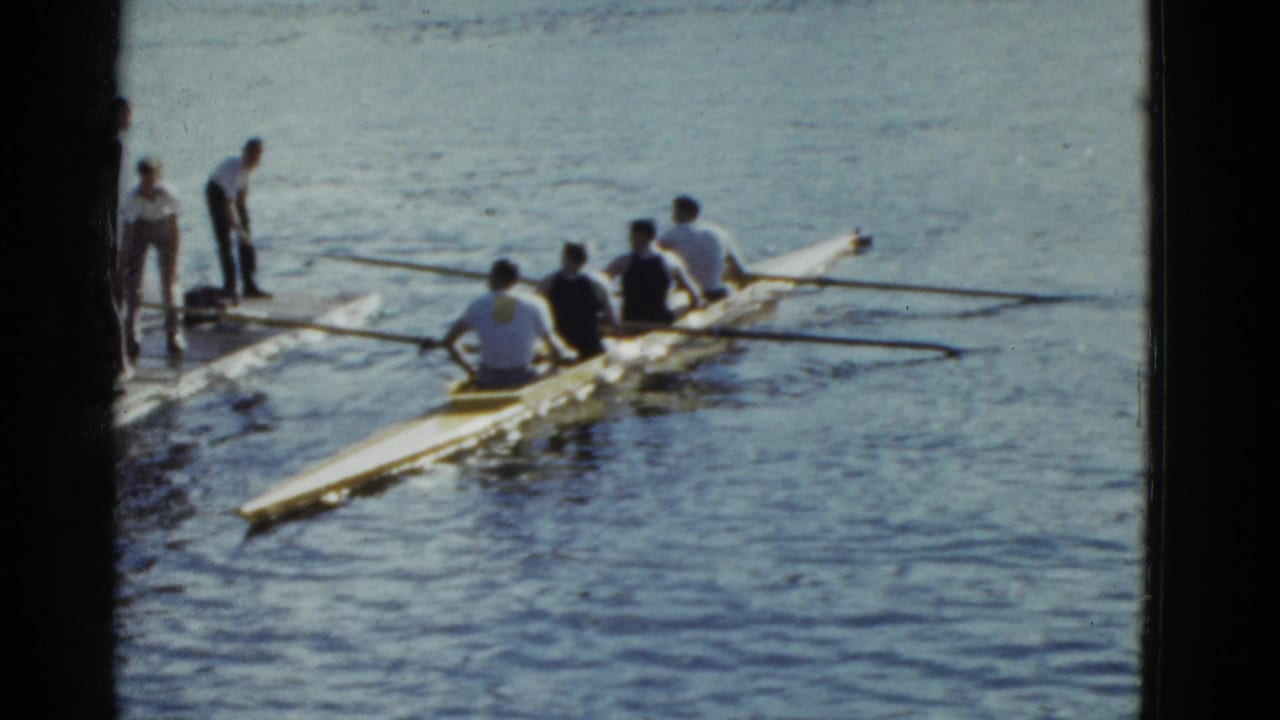 Rowing crew in a shell near a dock with oars stretched across the water.
