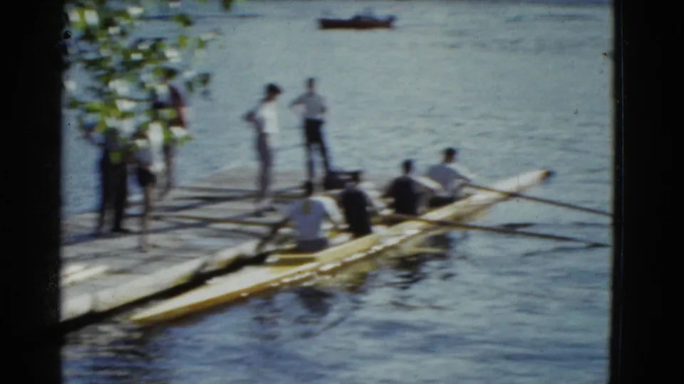 1975 Boston waterfront frame showing a crew shell beside the dock, nearby helpers, tree leaves, and a distant small boat.