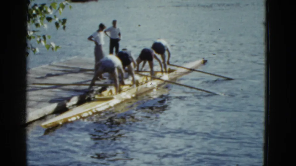 Adult rowers in 1975 Boston appear to adjust seats or oars on a yellow shell beside a wooden dock.