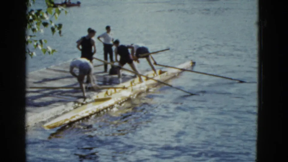1975 Boston home-movie still with men preparing a yellow rowing shell beside the dock as oars stretch outward.