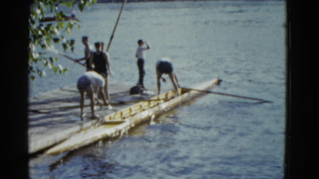 Young adults on a Boston river dock adjust a yellow rowing shell and oars in bright 1975 home-movie footage.