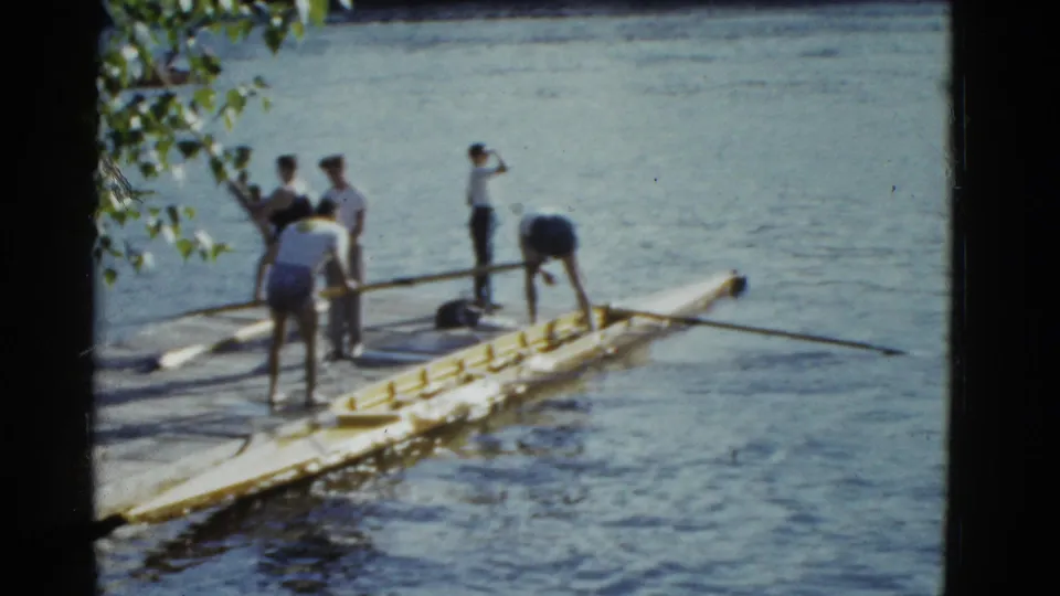 1975 Boston riverfront home-movie frame with young adults standing around a yellow rowing shell before departure.