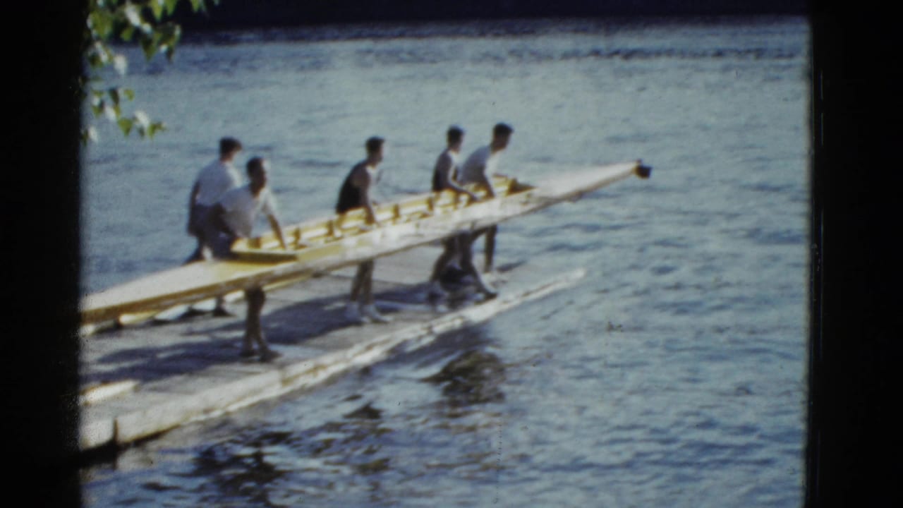 In 1975 Boston home-movie footage, young men gather around a long wooden rowing shell beside calm water and leafy branches.